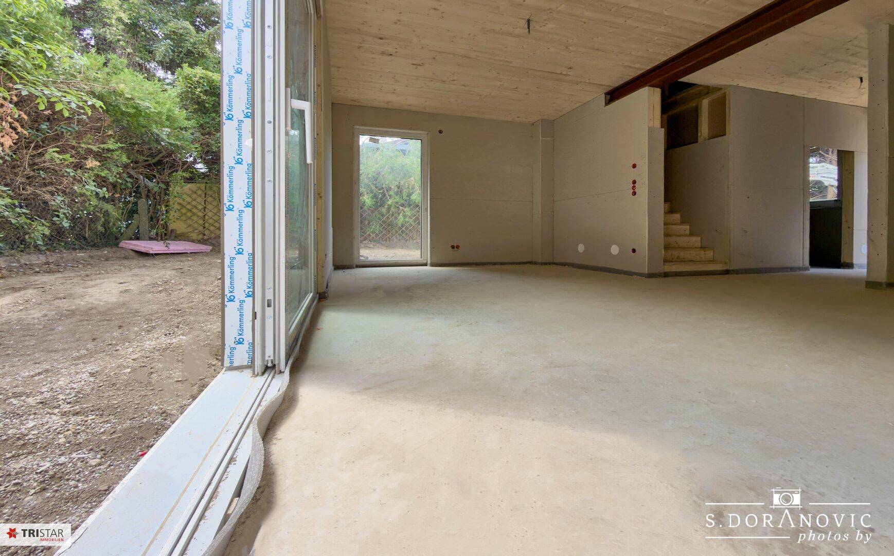 Geräumiger Rohbau-Innenraum mit Holzbalkendecke, großen Schiebetüren und Blick ins Grüne.