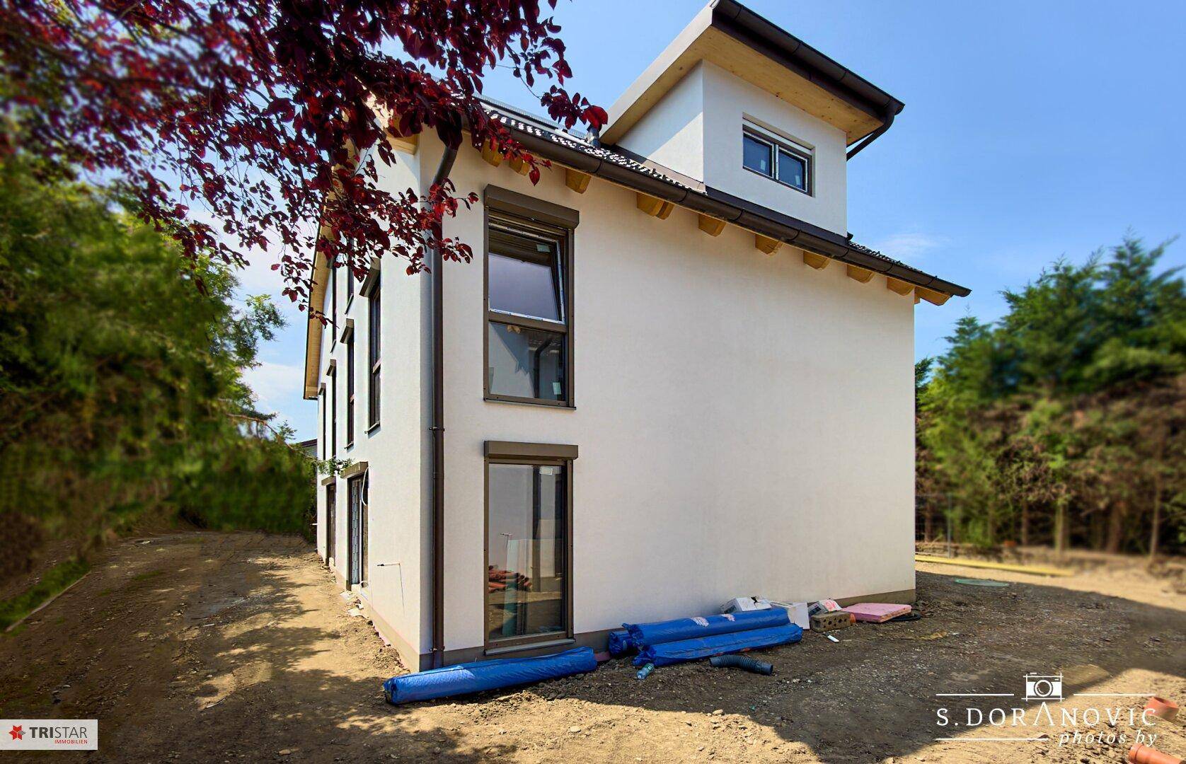 Seitenansicht eines modernen Neubaus mit weißer Fassade, Rollläden und Bauarbeiten im Gartenbereich.