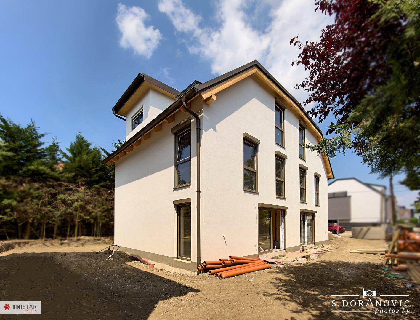 Modernes Einfamilienhaus im Bau mit weißer Fassade, dunklen Fenstern und umgebender Natur.