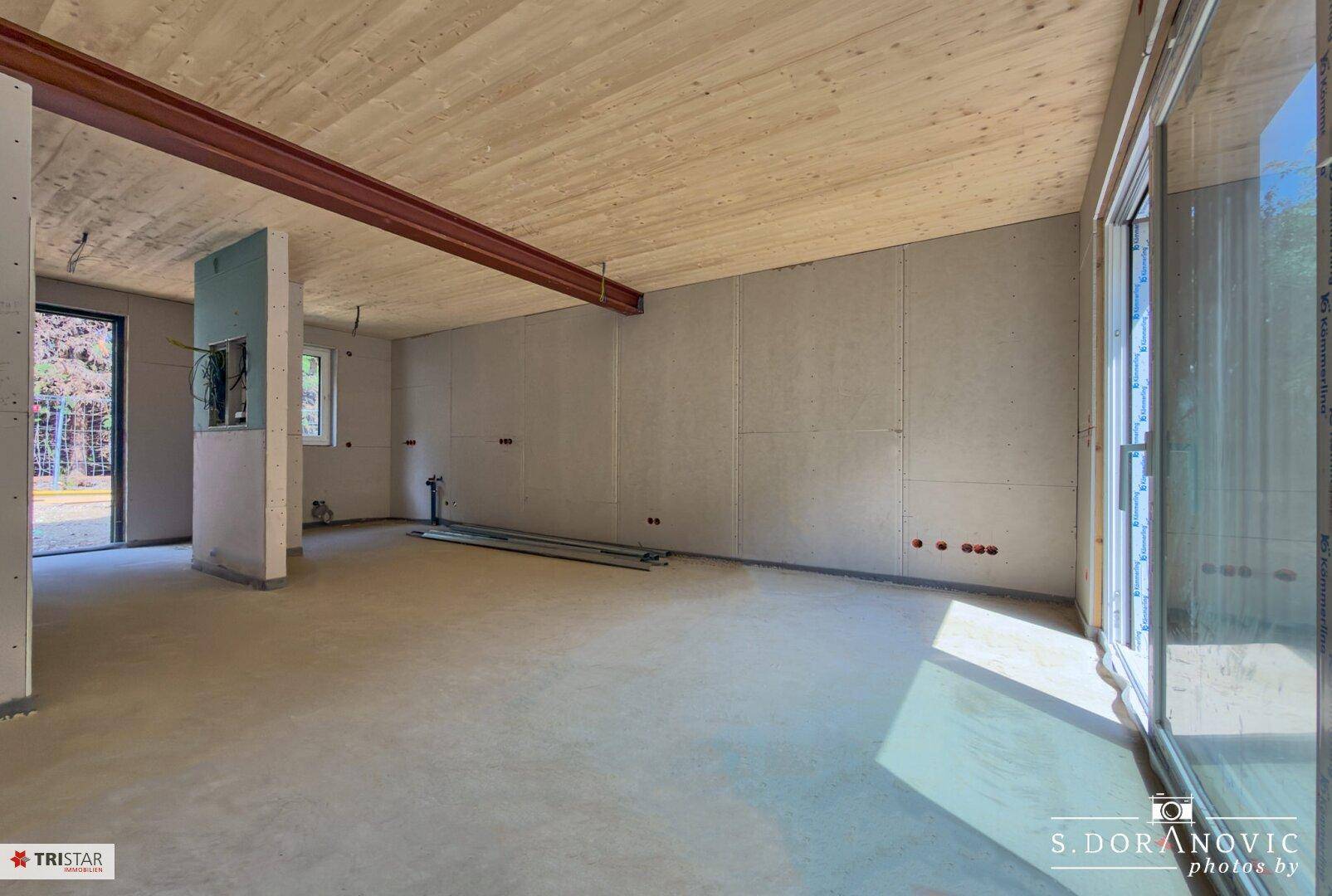Weitläufiger Rohbau-Wohnbereich mit Holzbalkendecke und viel Tageslicht durch große Fenster.