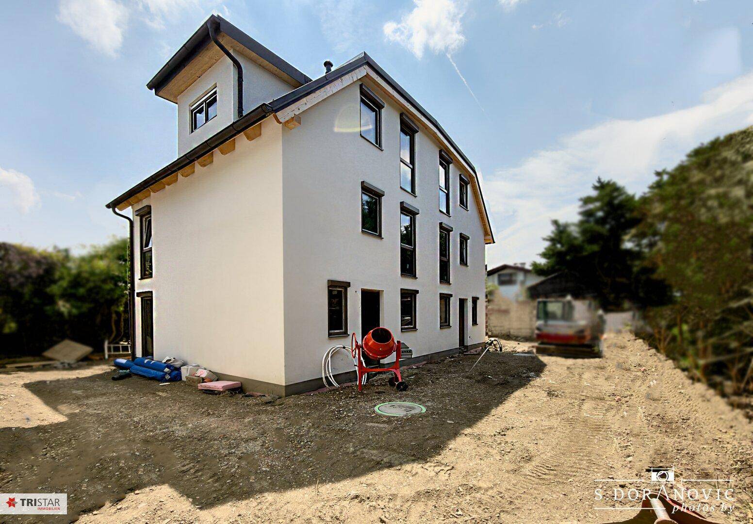 Seitenansicht des Neubaus mit weißer Fassade, dunklen Fenstern und Baustellenumgebung.