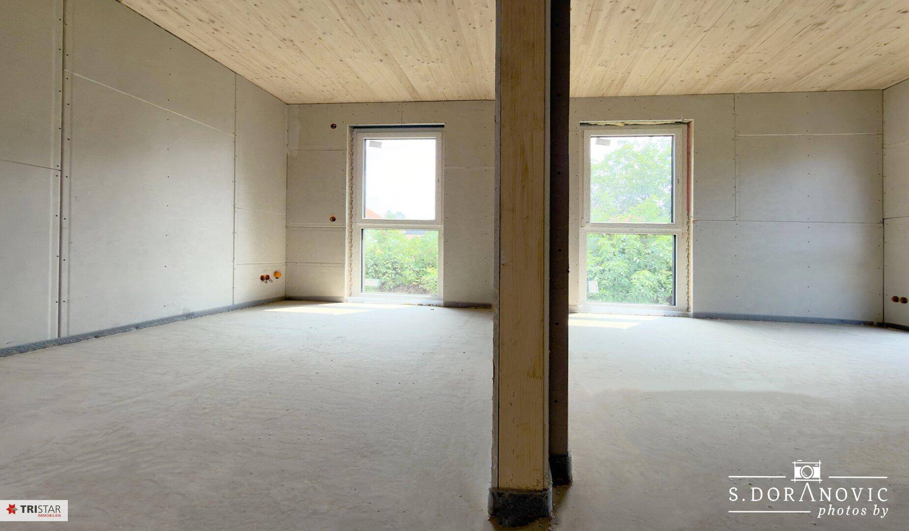 Heller, unfertiger Raum mit Betonboden, zwei großen Fenstern und Holzdeckenverkleidung.