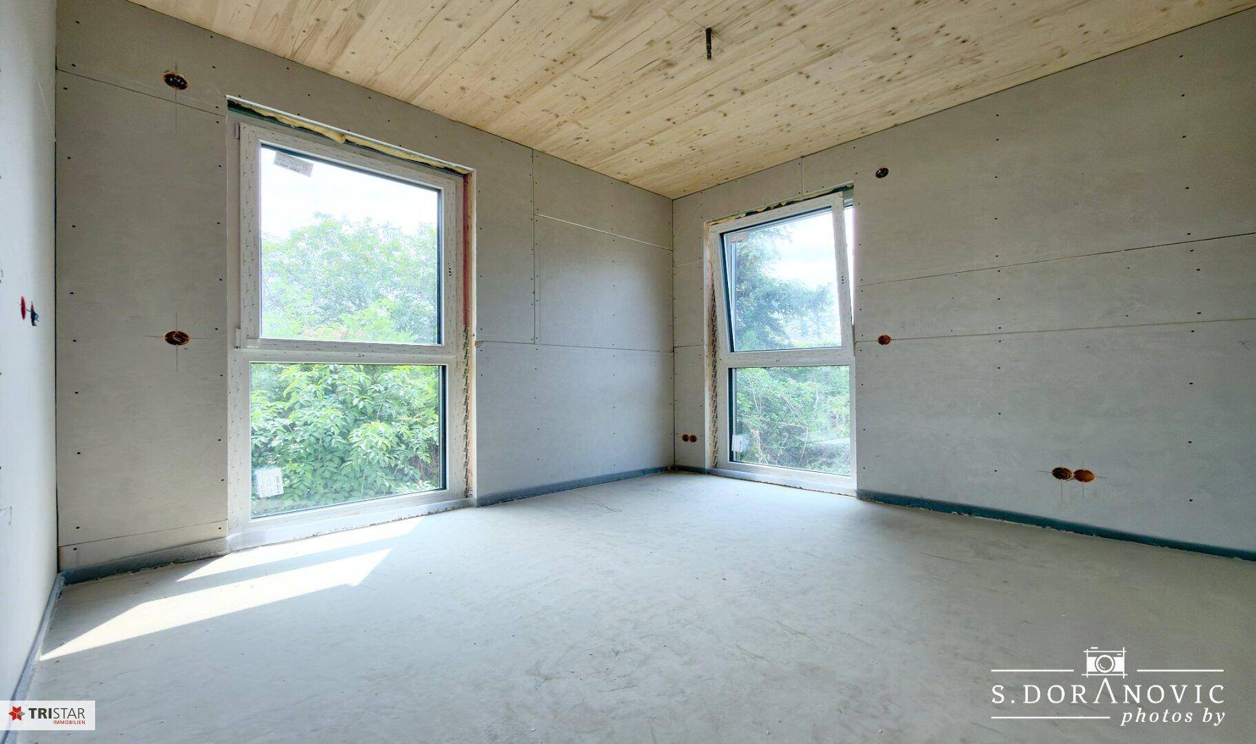 Heller, unfertiger Raum mit Betonboden, zwei großen Fenstern und Holzdeckenverkleidung.