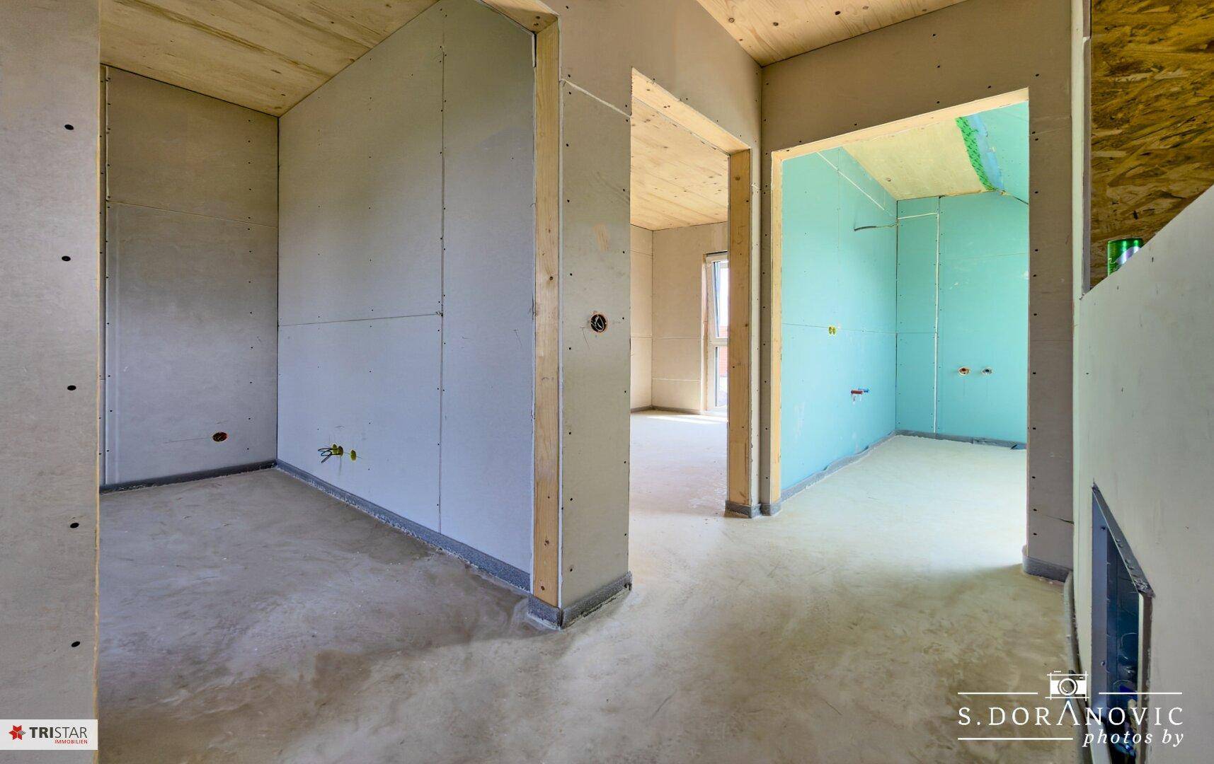 Unfertiger Flurbereich mit Betonboden, Holzdecken und Durchgängen zu weiteren Räumen.