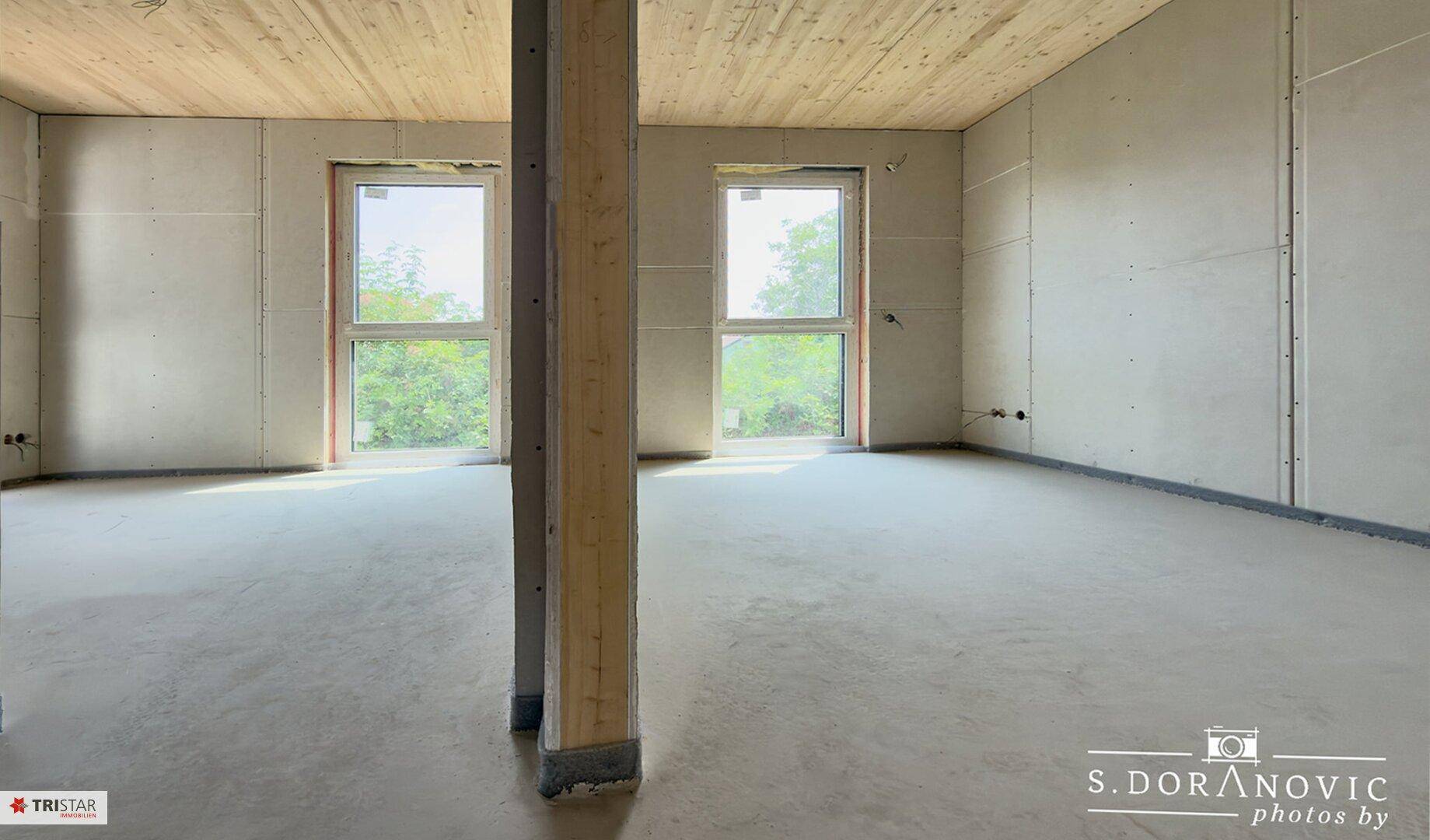 Unfertiger Innenraum mit Betonboden, großen Fenstern und Holzdeckenverkleidung, bereit für den Ausbau.