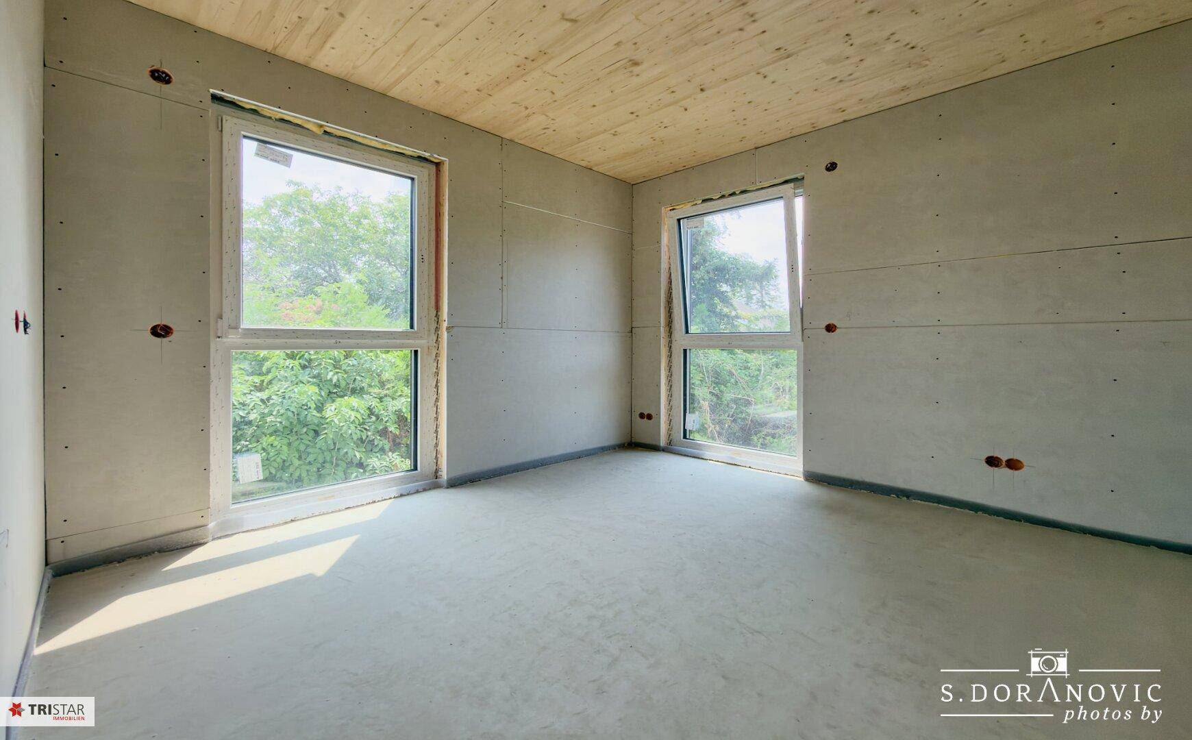 Heller, unfertiger Raum mit Betonboden, zwei großen Fenstern und Holzdeckenverkleidung.