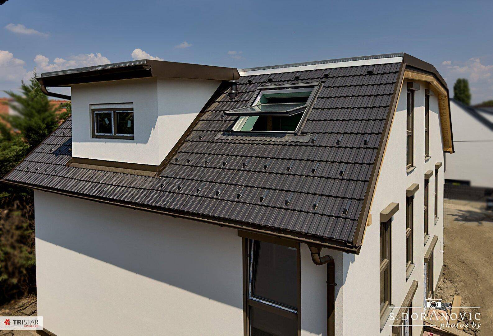 Dachansicht eines Neubaus mit dunklen Ziegeln, Dachfenster und Gaube unter klarem Himmel.