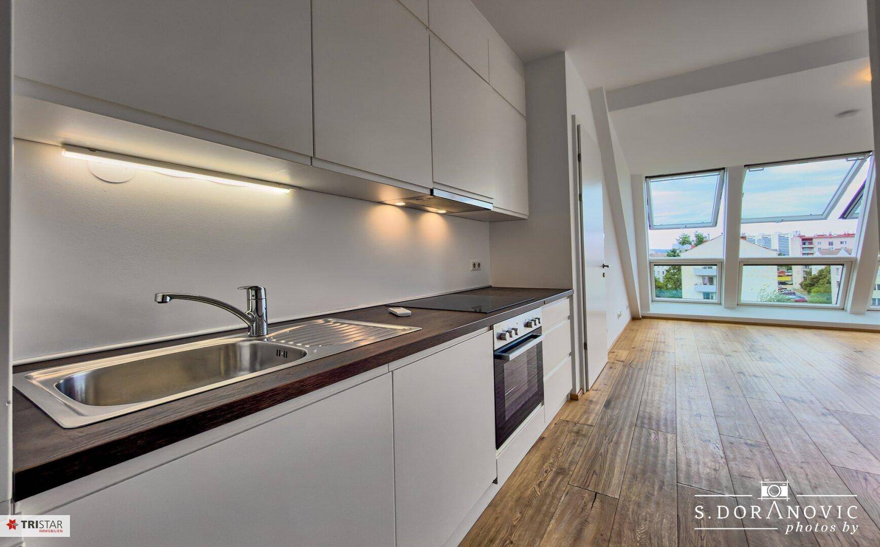 Moderne Einbauküche mit weißen Schränken und dunkler Arbeitsplatte, angrenzend an einen hellen Wohnbereich.