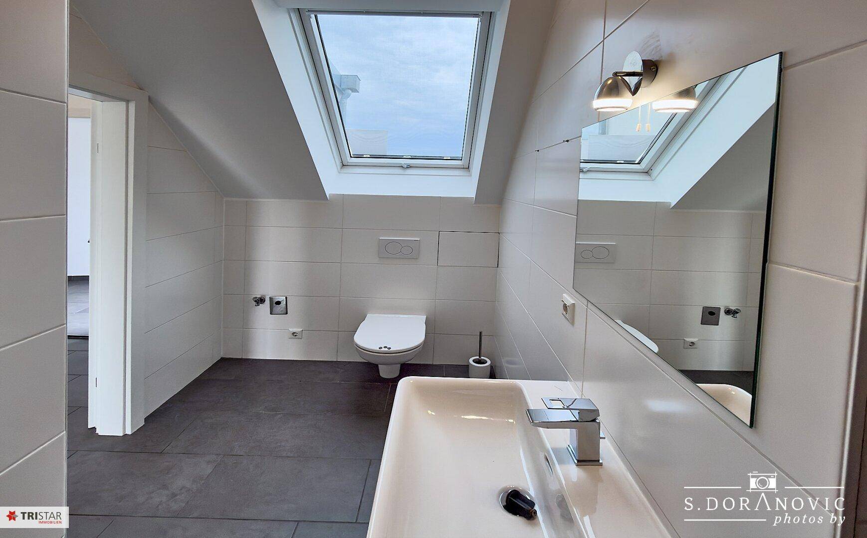 Modernes Badezimmer mit weißen Fliesen, Waschbecken, Toilette und Dachfenster für Tageslicht.