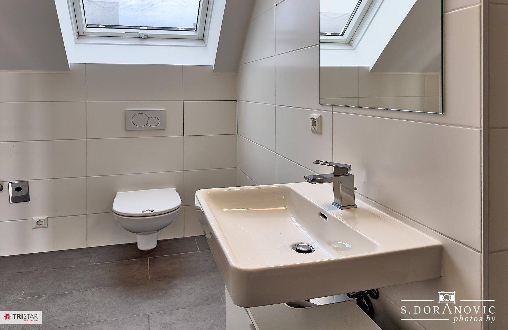 Helles Badezimmer mit modernem Waschbecken, Wand-WC und großem Spiegel unter einem Dachfenster.