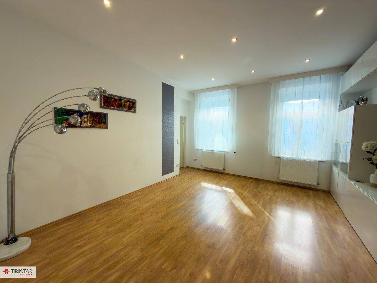 Geräumiges Wohnzimmer mit hellem Laminatboden und großen Fenstern, die viel Licht hereinlassen.