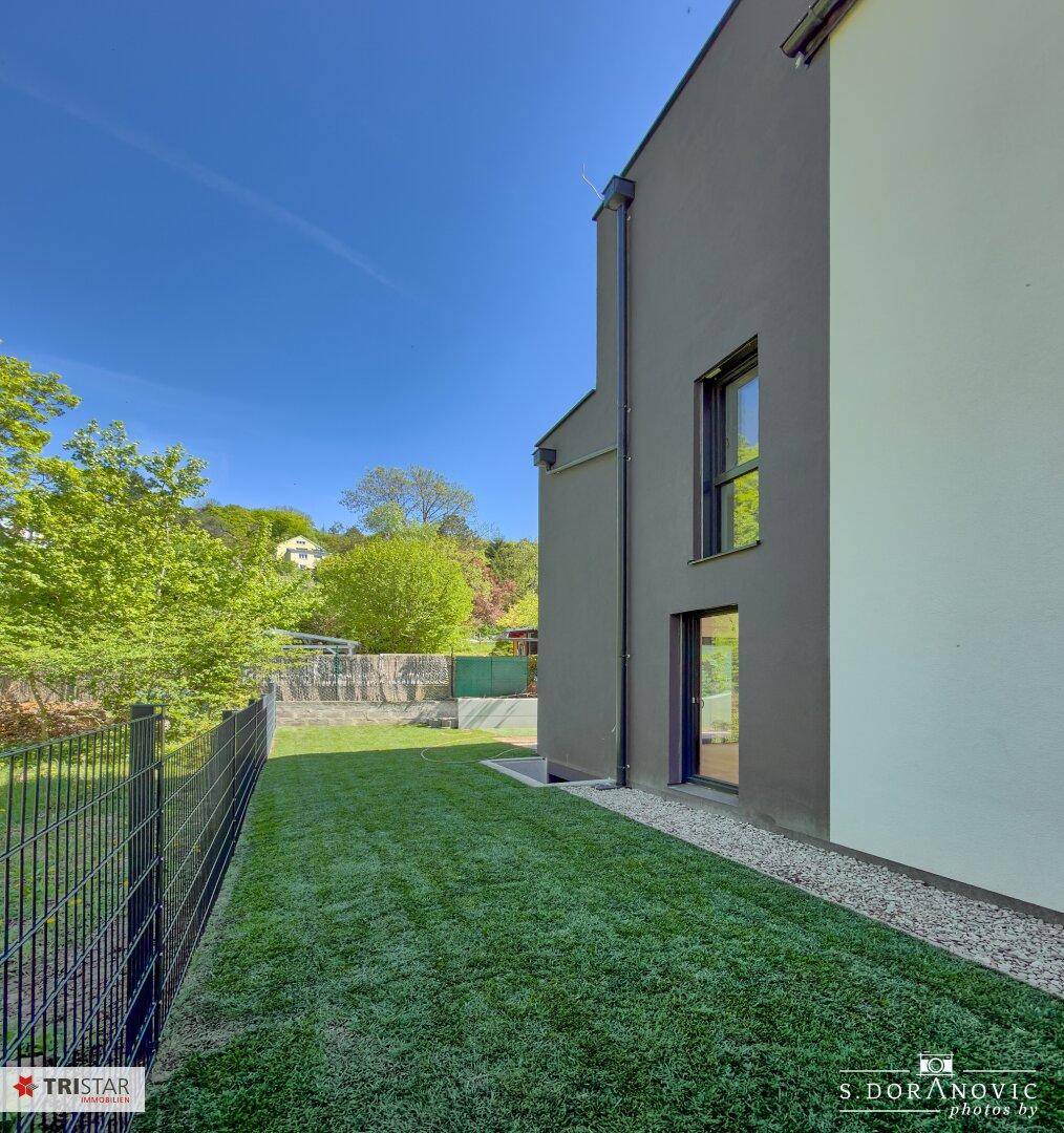 Schmaler, gepflegter Rasenstreifen entlang der Hausfassade, begrenzt durch einen Metallzaun.