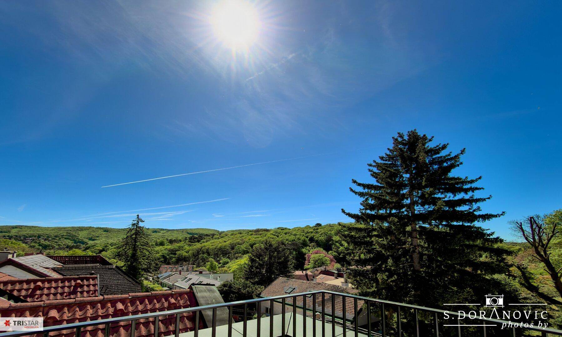 Weitläufiger Blick von einem Balkon über rote Dächer und grüne Hügel unter einem strahlend blauen Himmel.