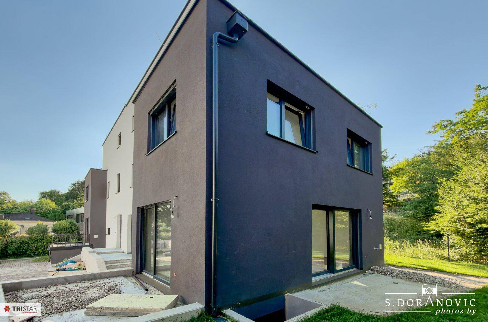 Eckansicht eines modernen Neubaus mit dunkler Fassade und großen Fensterfronten, umgeben von Baustellenelementen.