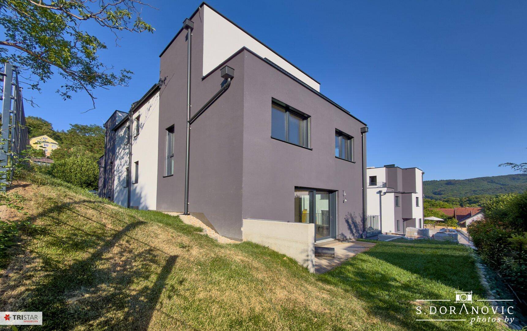 Seitenansicht eines modernen Neubaus mit grauer Fassade und angrenzendem grünen Hang.
