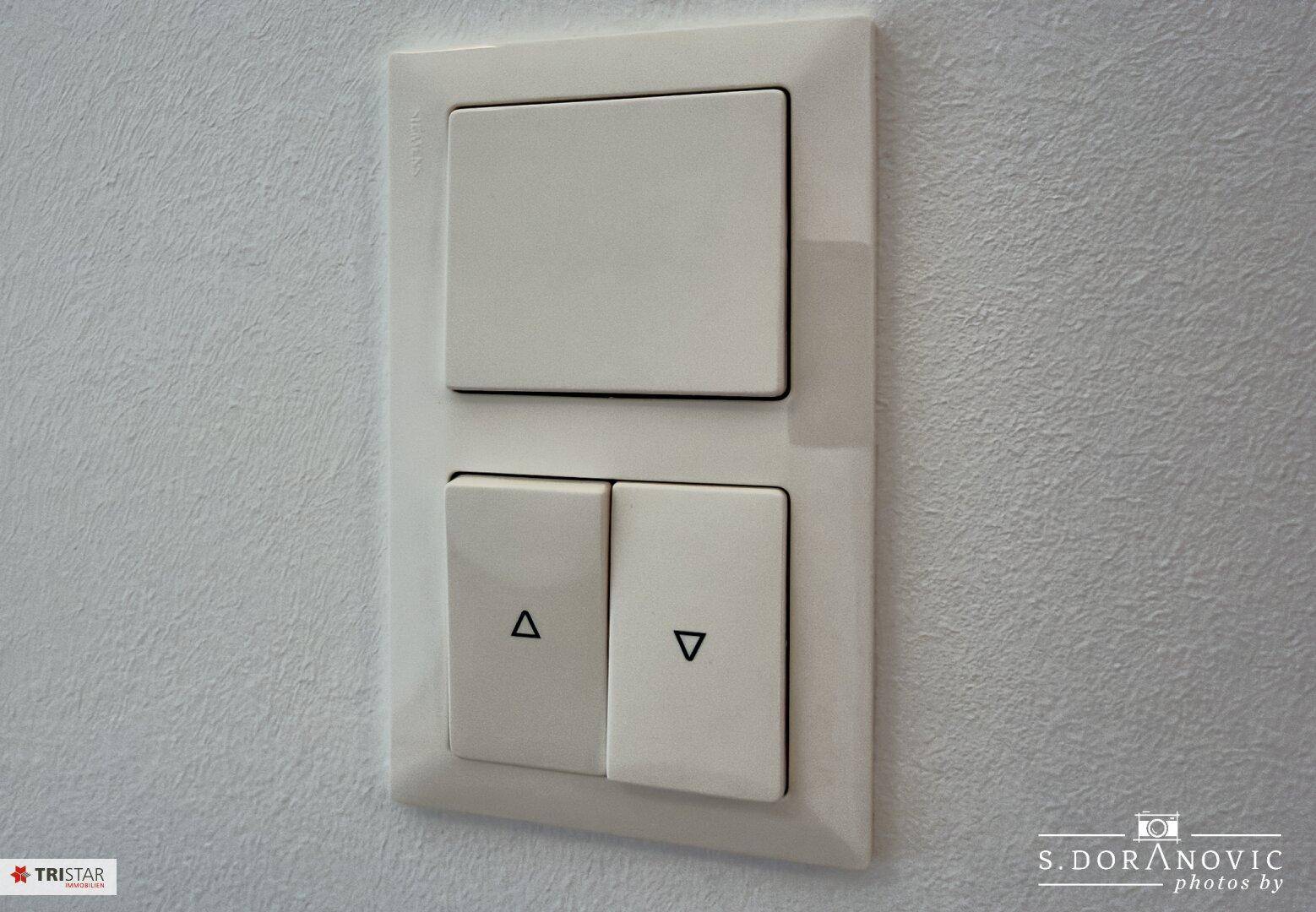 Nahaufnahme eines modernen Lichtschalters und Rollladenbedienfelds an einer weißen Wand.