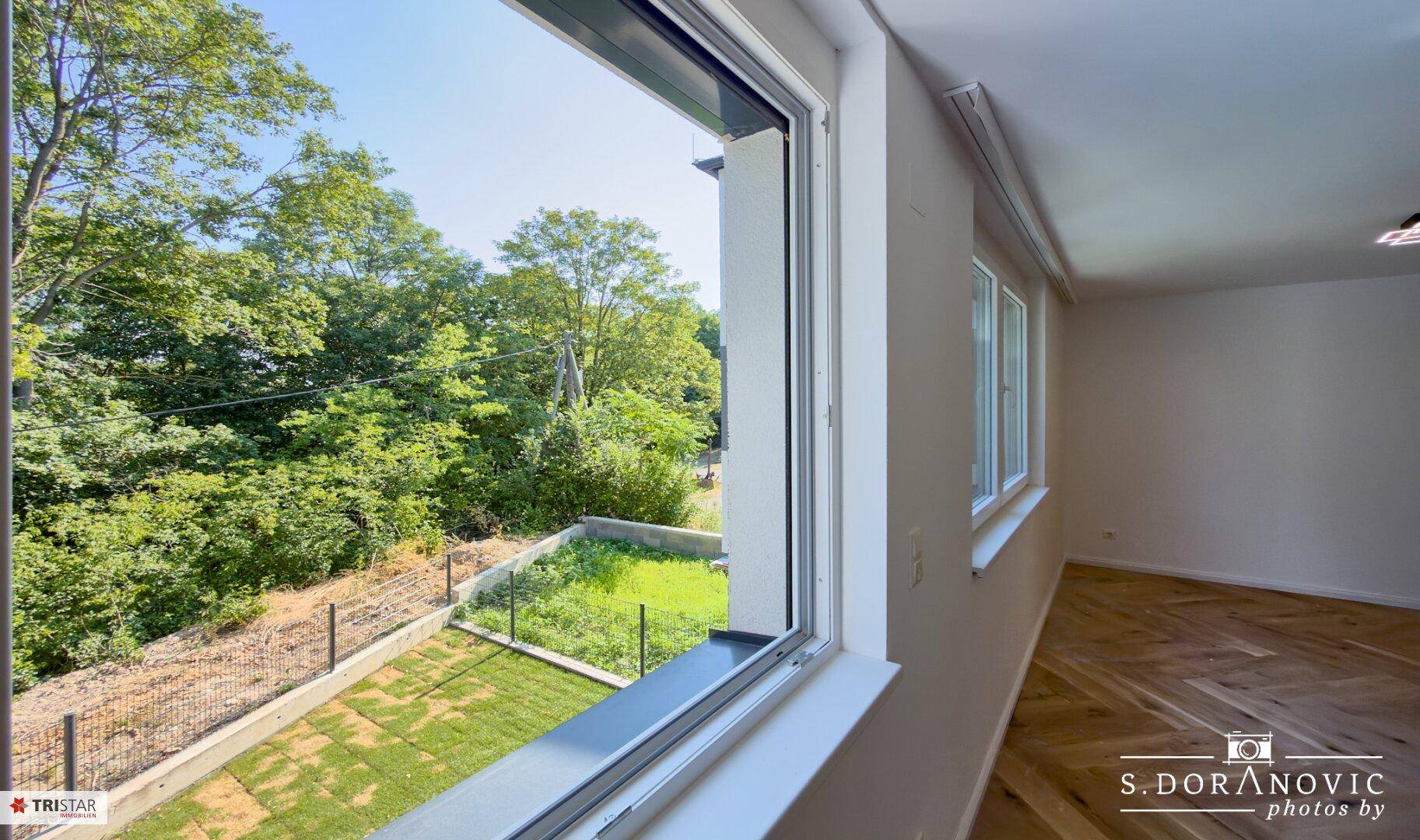 Helles Zimmer mit Fischgrätparkett und großem Fenster, das einen weiten Blick ins Grüne bietet.