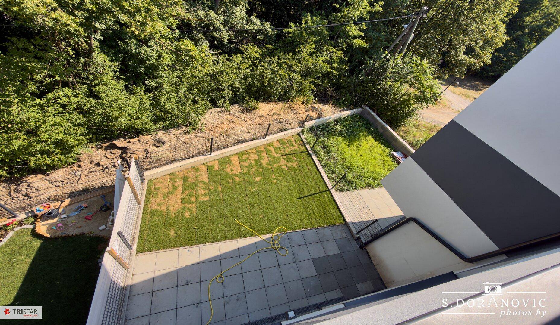 Blick von oben auf den Gartenbereich mit Terrasse und angrenzendem Grün, umgeben von Bäumen.