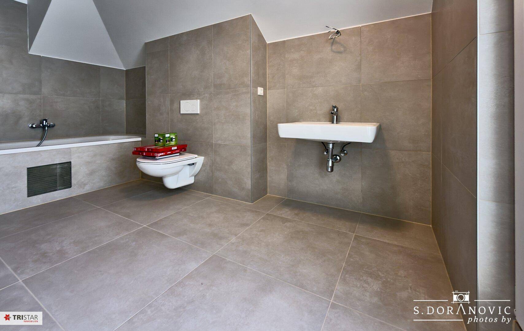 Modernes Badezimmer mit grauen Großformatfliesen, wandhängendem WC, Waschtisch und Badewanne.