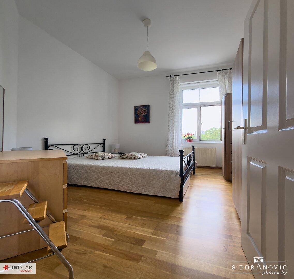 Schlafzimmer mit Holzboden, Doppelbett und Kleiderschrank, hell durch großes Fenster.