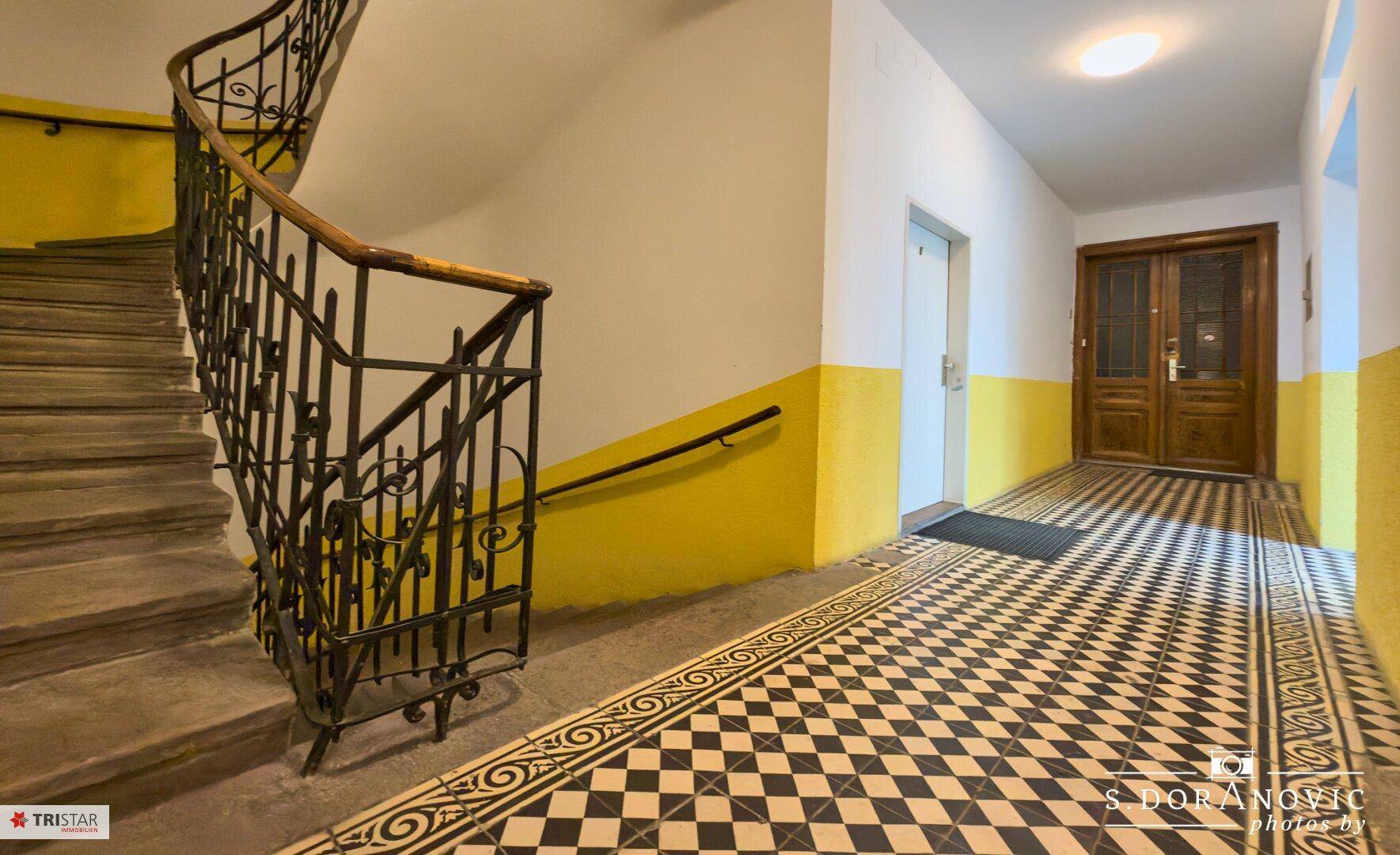 Eingangsbereich mit historischem Treppenhaus, gemusterten Fliesen und gelben Wänden.