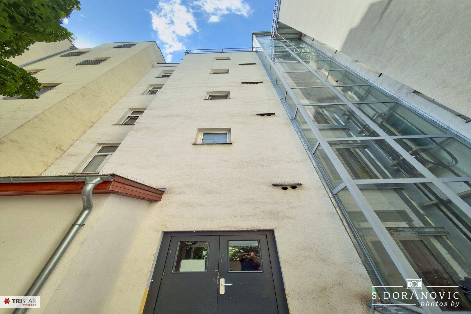 Blick auf die Gebäudefassade mit einem modernen Glasaufzug, der die oberen Stockwerke erschließt.