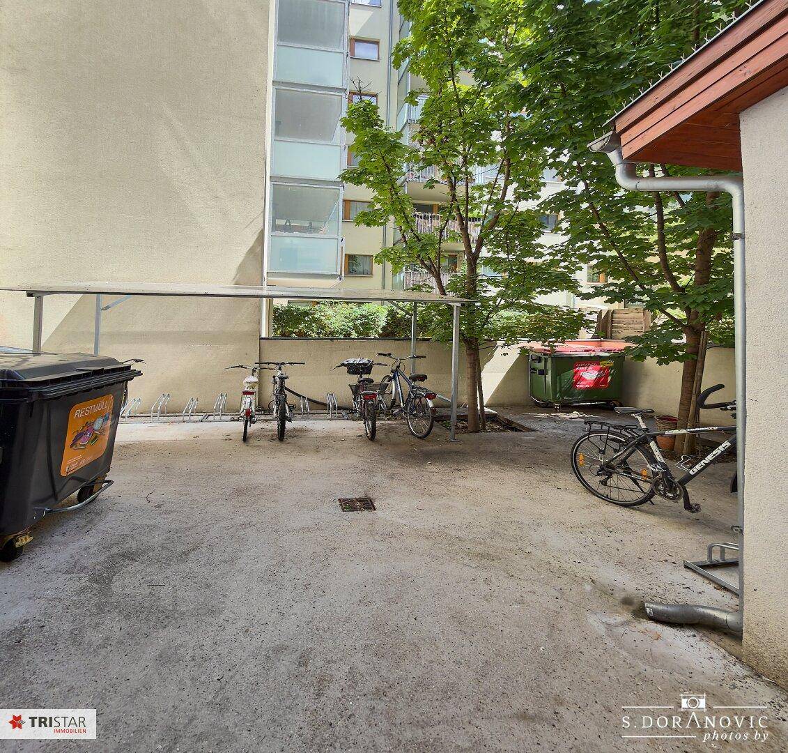 Außenbereich mit Fahrradabstellplätzen und Müllcontainern, umgeben von Bäumen.