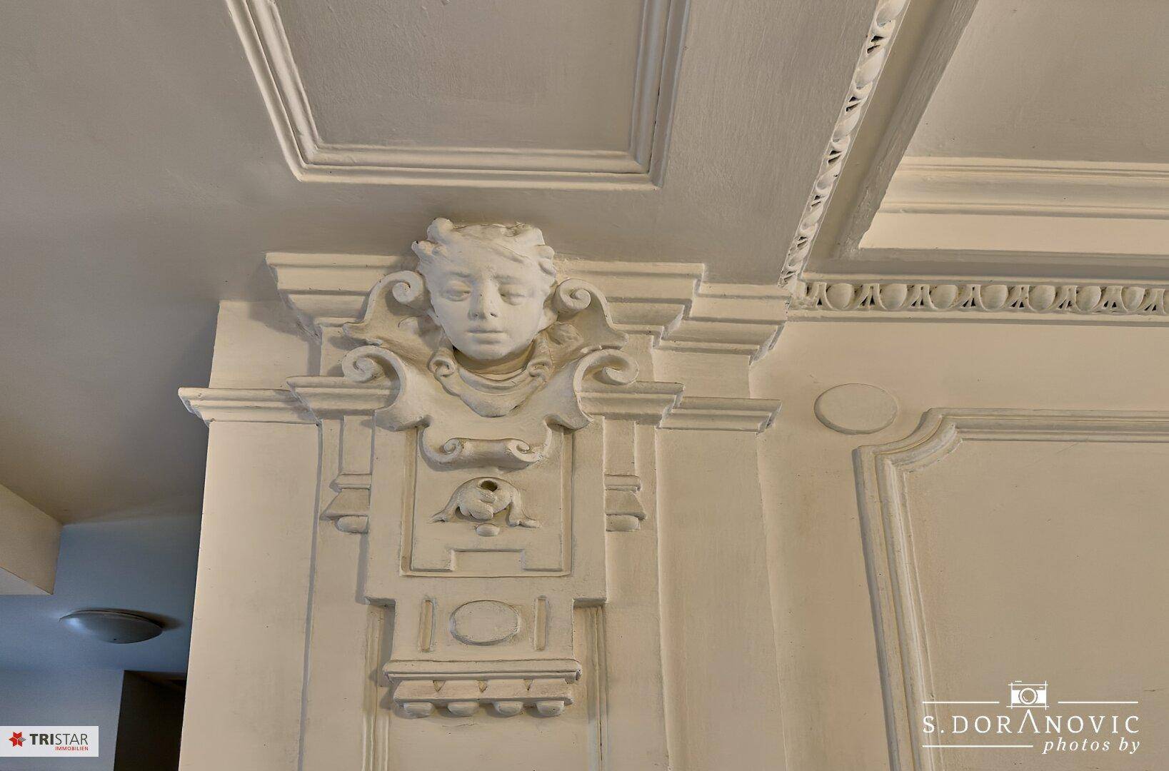 Detailaufnahme einer kunstvollen Stuckverzierung an Wand und Decke mit einem antiken Gesichtsmotiv.