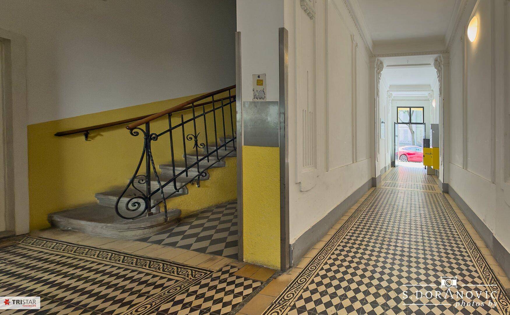 Historischer Flur mit Schachbrettmuster-Fliesenboden und einer Treppe mit gelb gestrichener Wand.