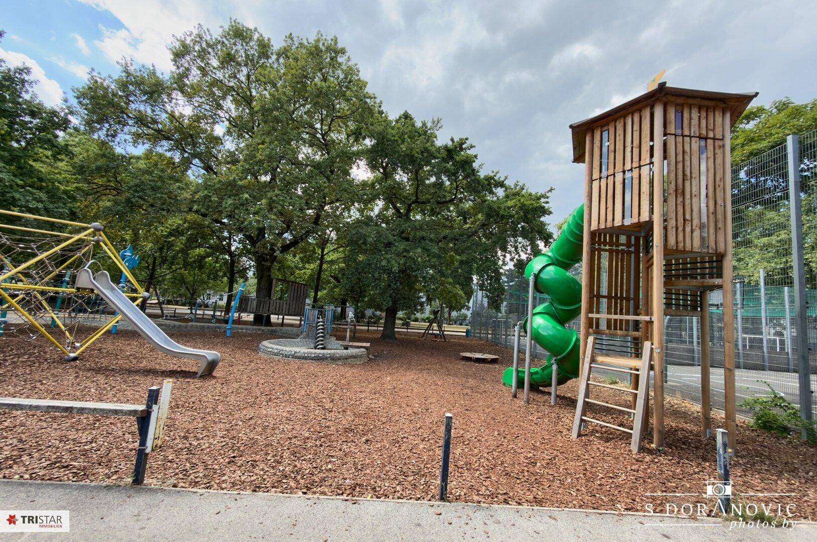 Weitläufiger Spielplatz mit Holzturm, grüner Rutsche und vielen Bäumen im Hintergrund.