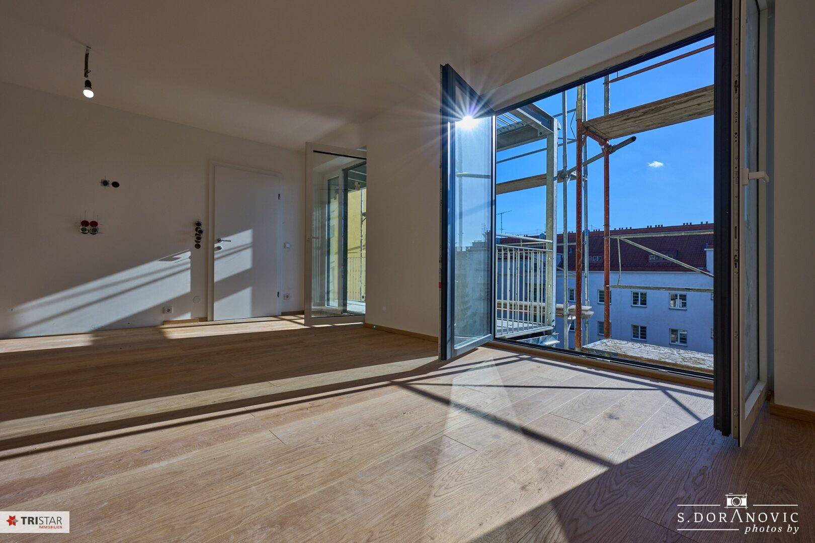 Heller Wohnbereich mit Holzboden und großen Fenstern, die viel Sonnenlicht hereinlassen.