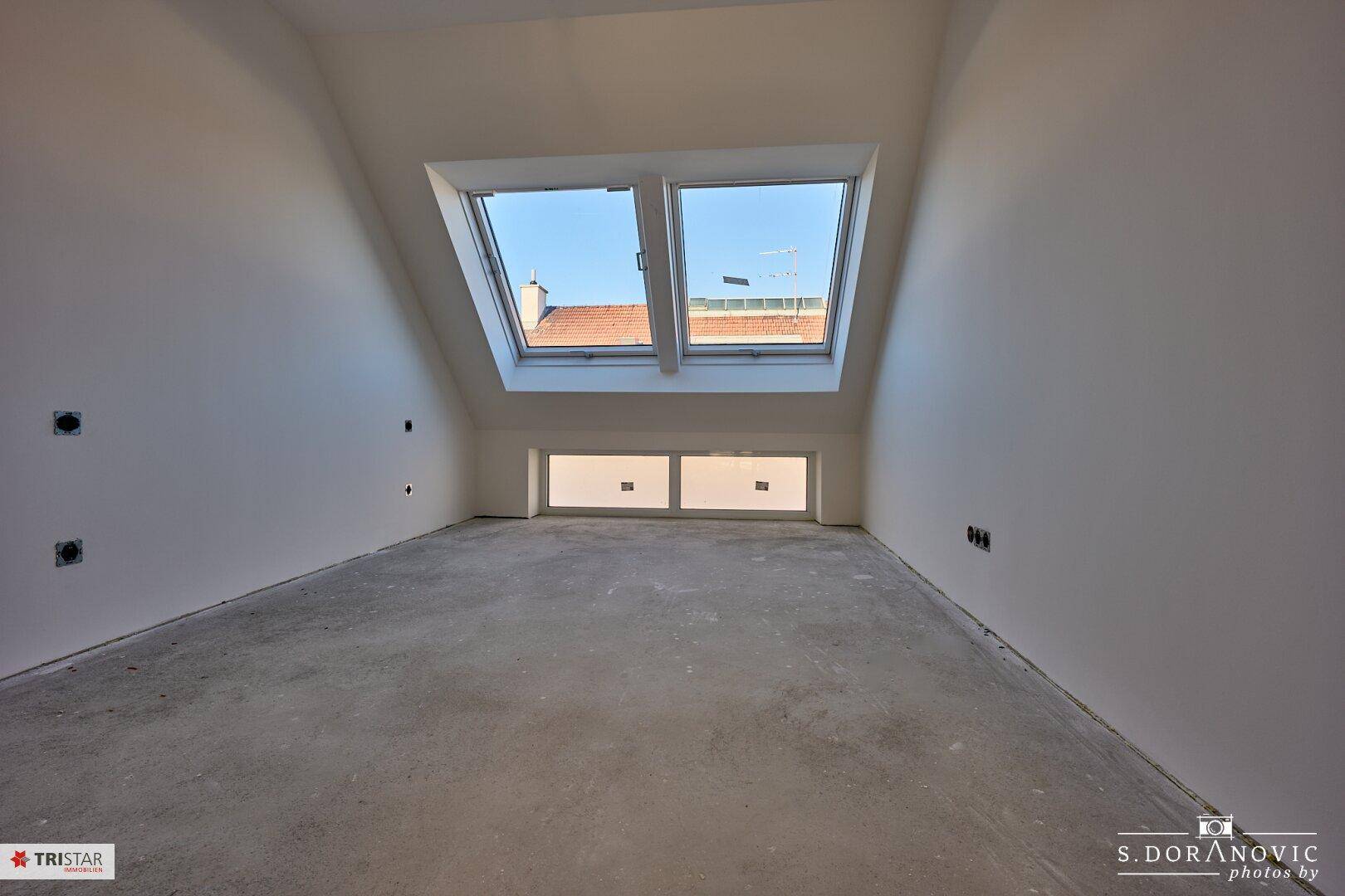Unfertiges Dachzimmer mit schrägen Wänden, zwei Dachfenstern und einem niedrigeren Fenster für zusätzliches Licht.