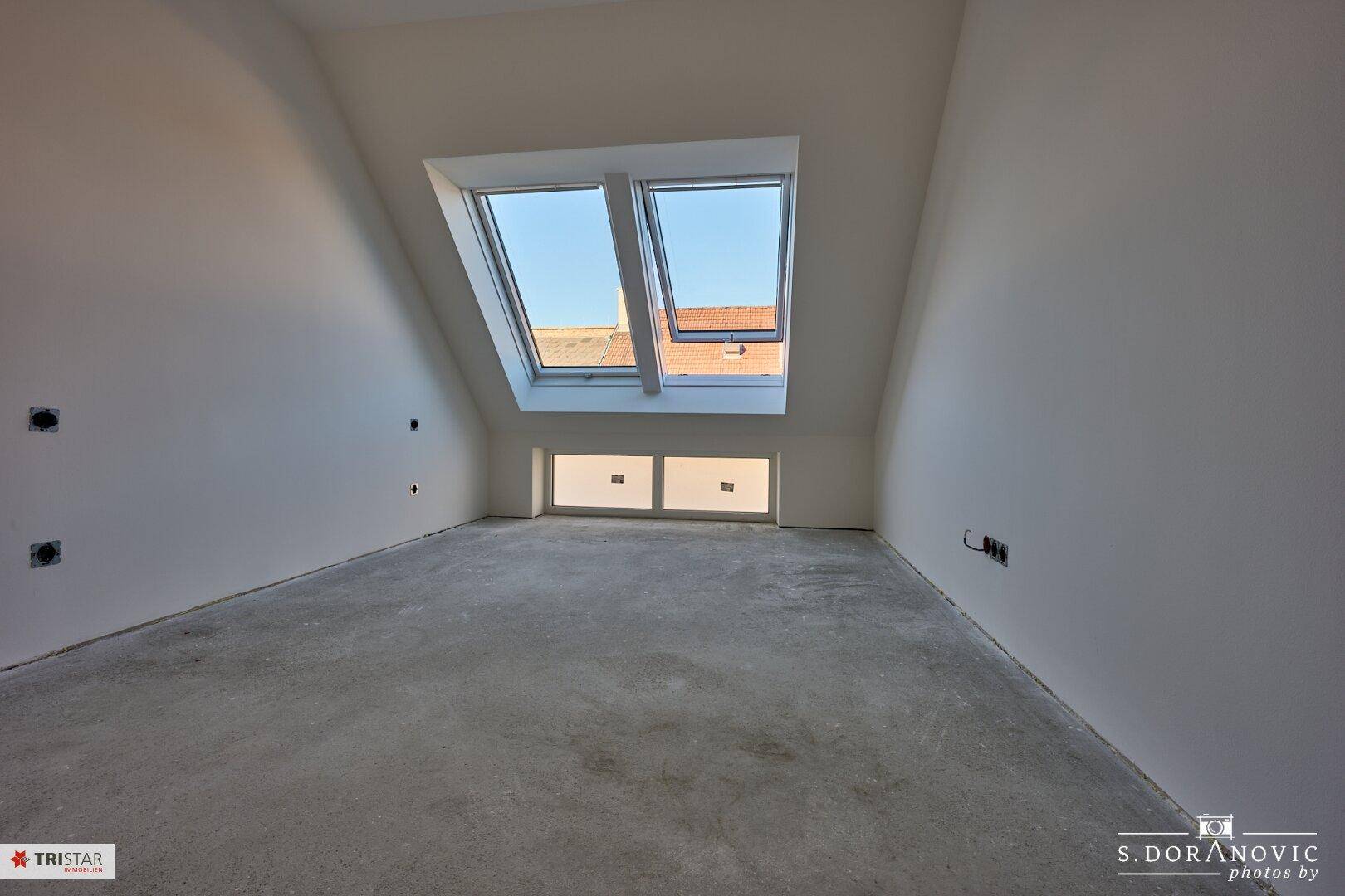 Unfertiges Dachzimmer mit schrägen Wänden, zwei Dachfenstern und einem niedrigeren Fenster für zusätzliches Licht.