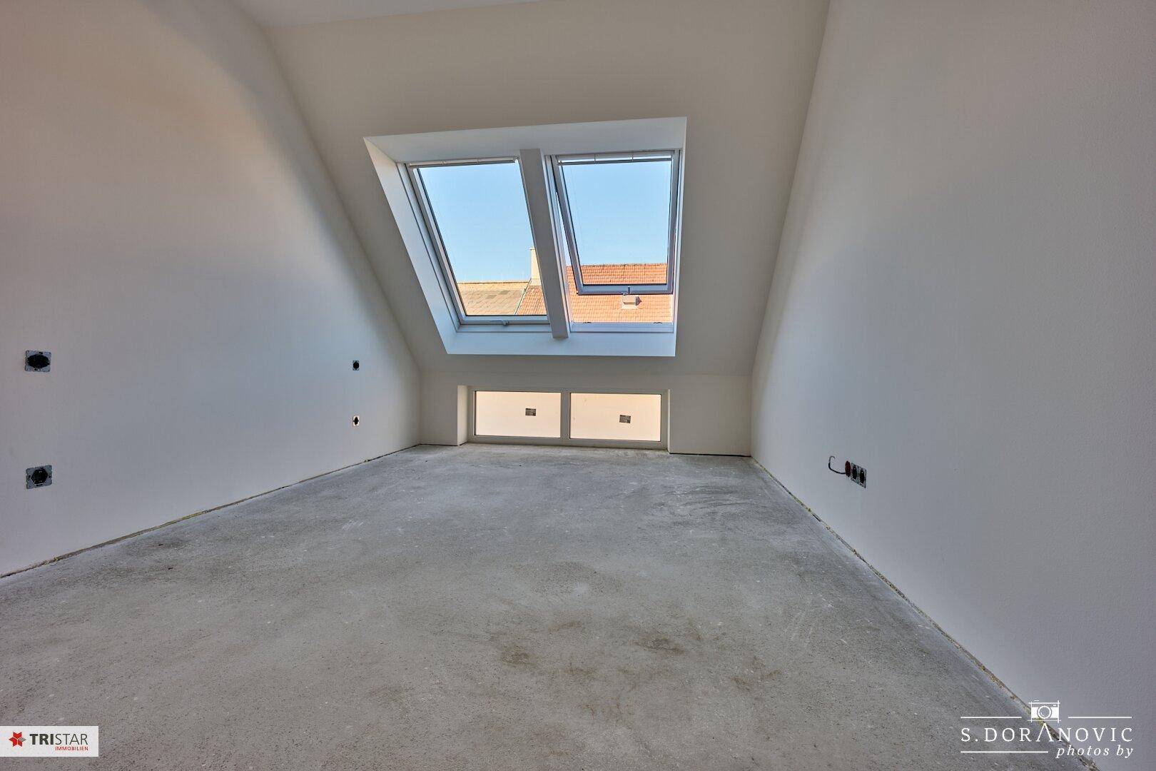 Unfertiges Dachzimmer mit schrägen Wänden, zwei Dachfenstern und einem niedrigeren Fenster für zusätzliches Licht.