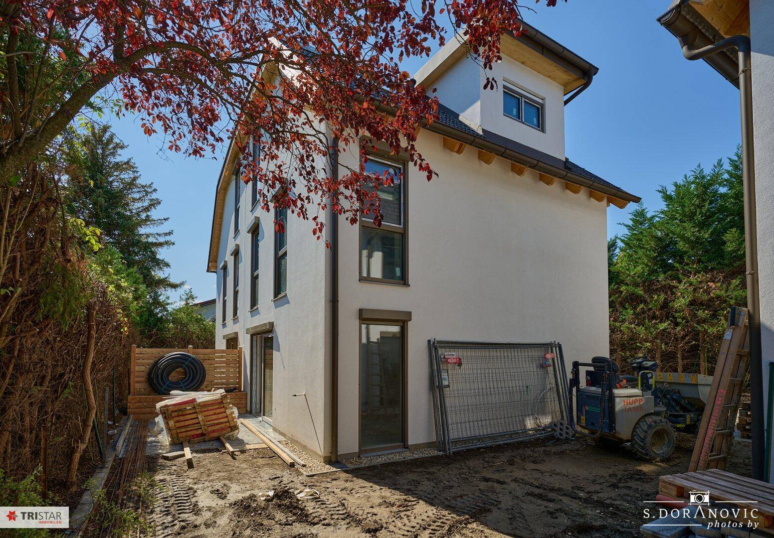 Neubau-Doppelhaushälfte von der Seite mit Baustellenelementen und einem Baum im Vordergrund.