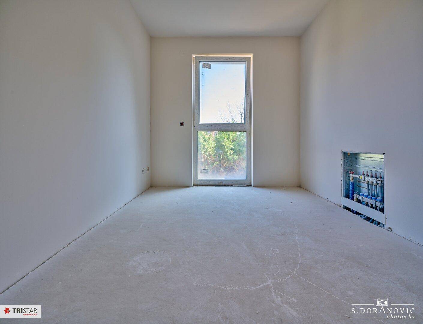Helles Schlafzimmer im Rohbau mit Fenster und sichtbaren Heizungsanschlüssen.