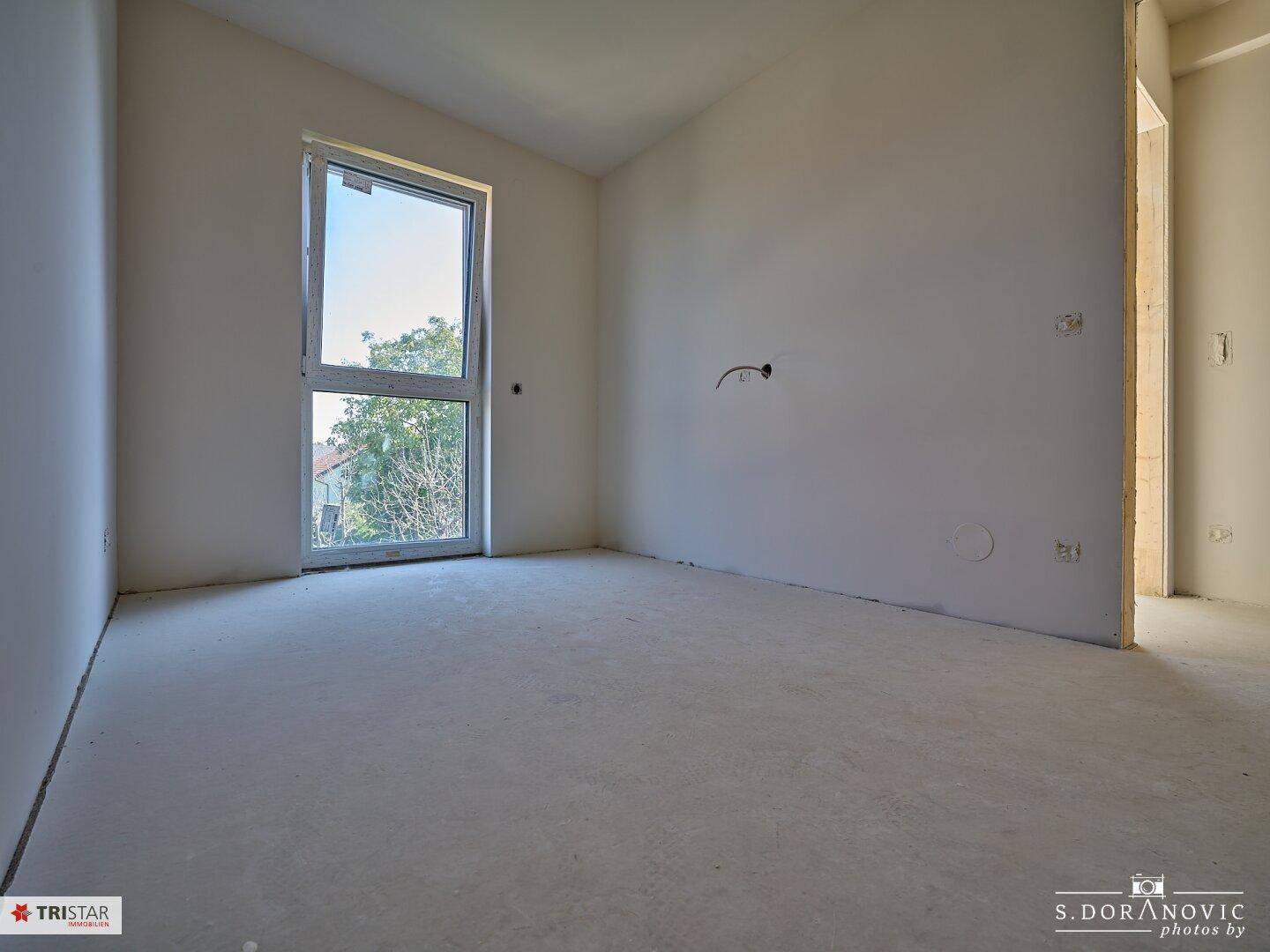 Helles Schlafzimmer im Rohbau mit großem Fenster und Blick auf die Umgebung.