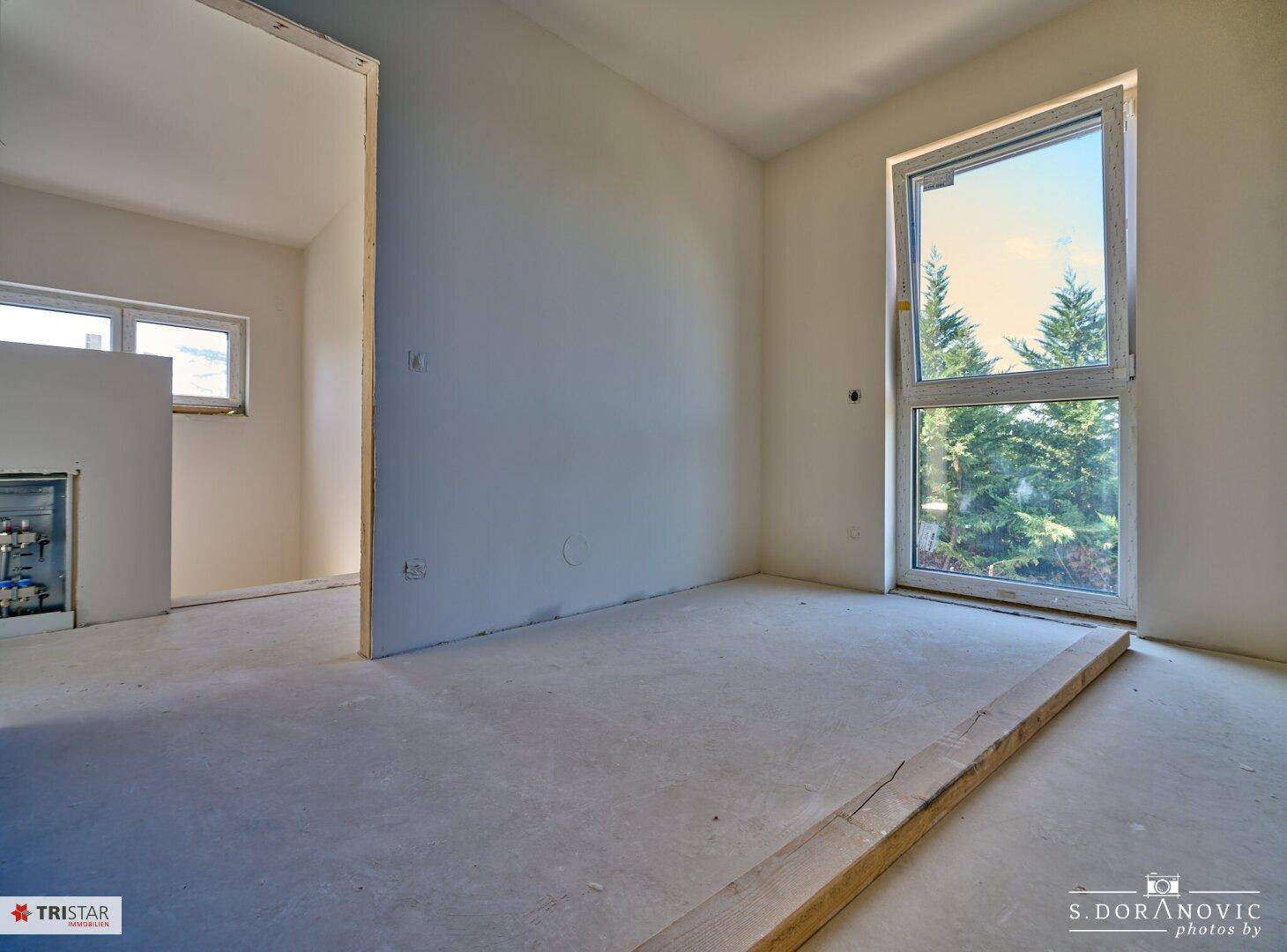 Helles Schlafzimmer im Rohbau mit großem Fenster und Blick auf die Bäume.
