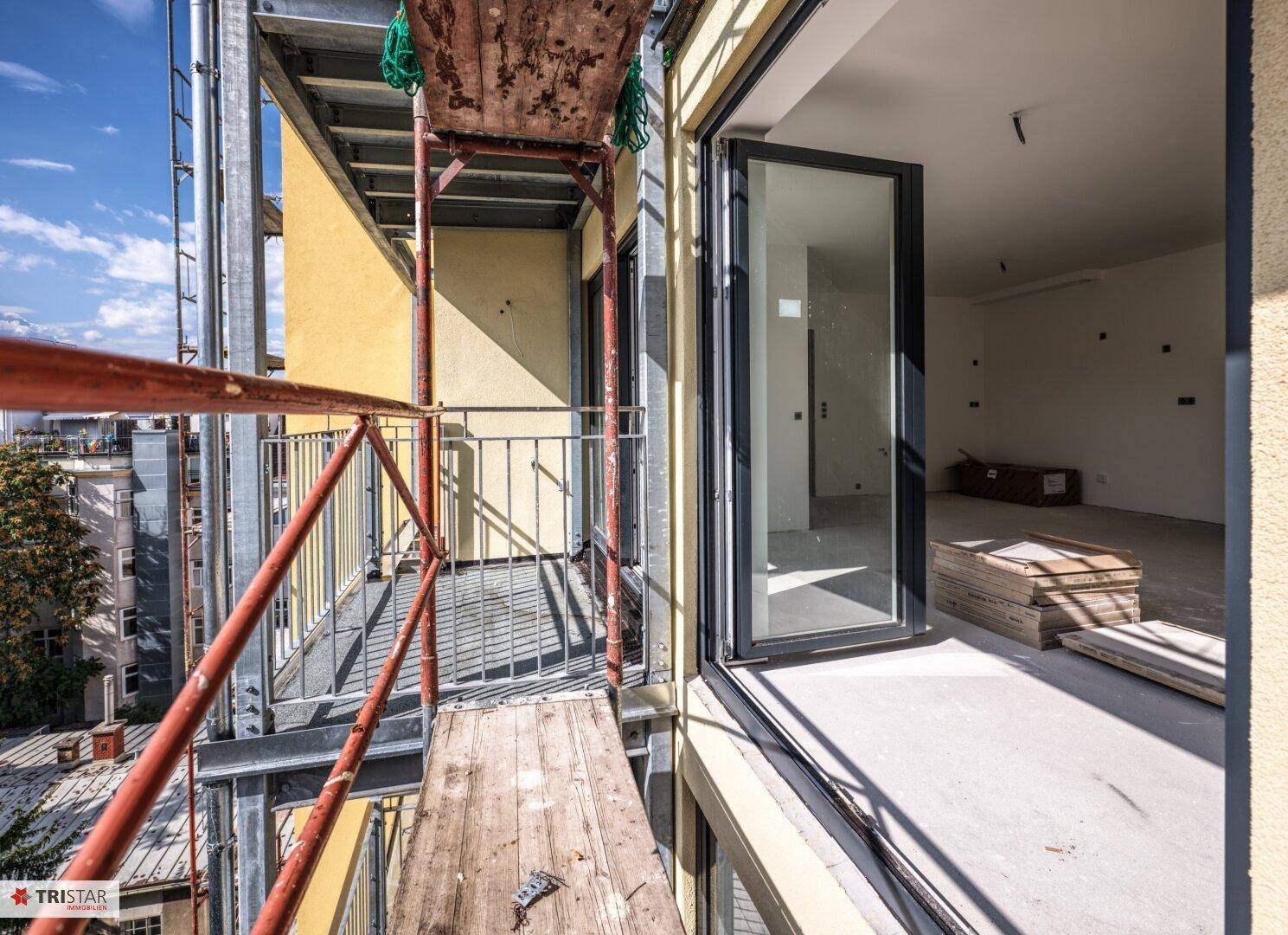 Blick vom Balkon in einen hellen, unfertigen Innenraum mit großen Fenstern und Betonboden.