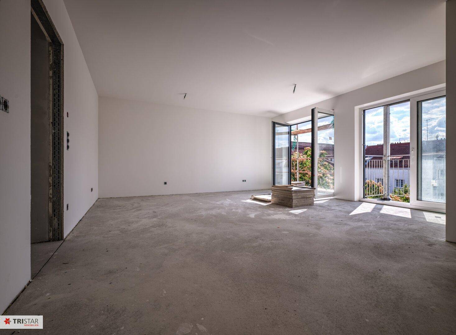 Großer, heller Wohnraum im Rohbauzustand mit Betonboden und großen Fensterfronten zum Balkon.
