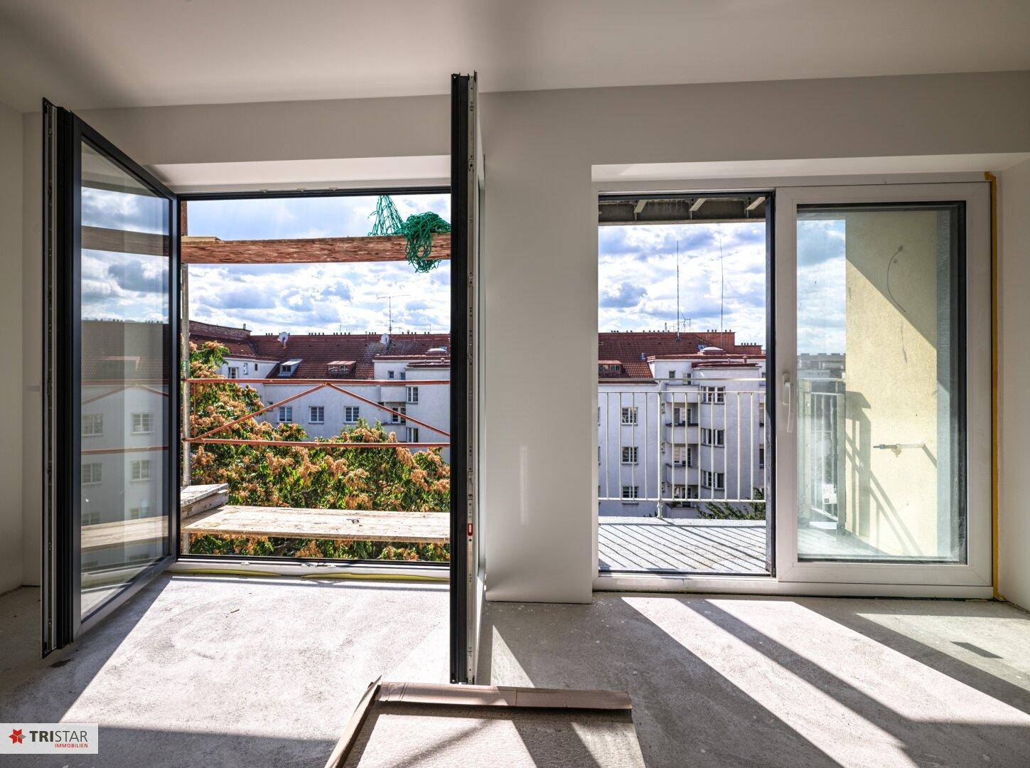 Lichtdurchfluteter Raum mit geöffneten Fenstern, Blick auf Bäume und umliegende Gebäude der Stadt.