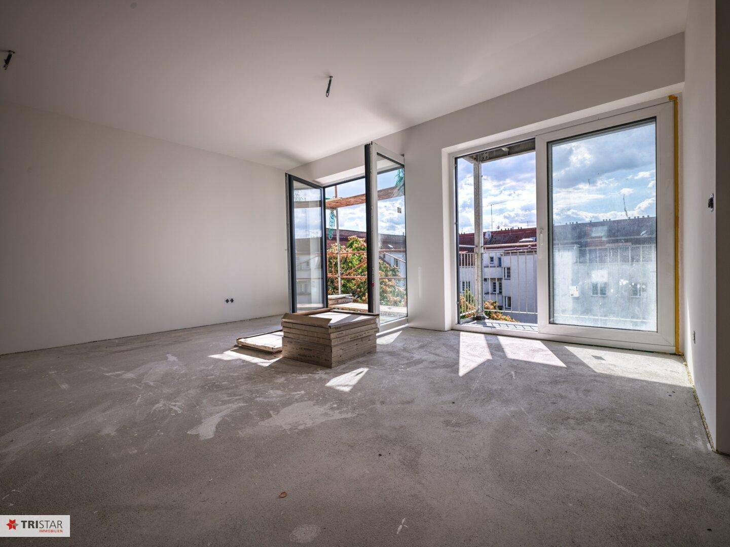 Heller, unfertiger Wohnraum mit Betonboden und großen Fenstern, die Zugang zu einem Balkon bieten.