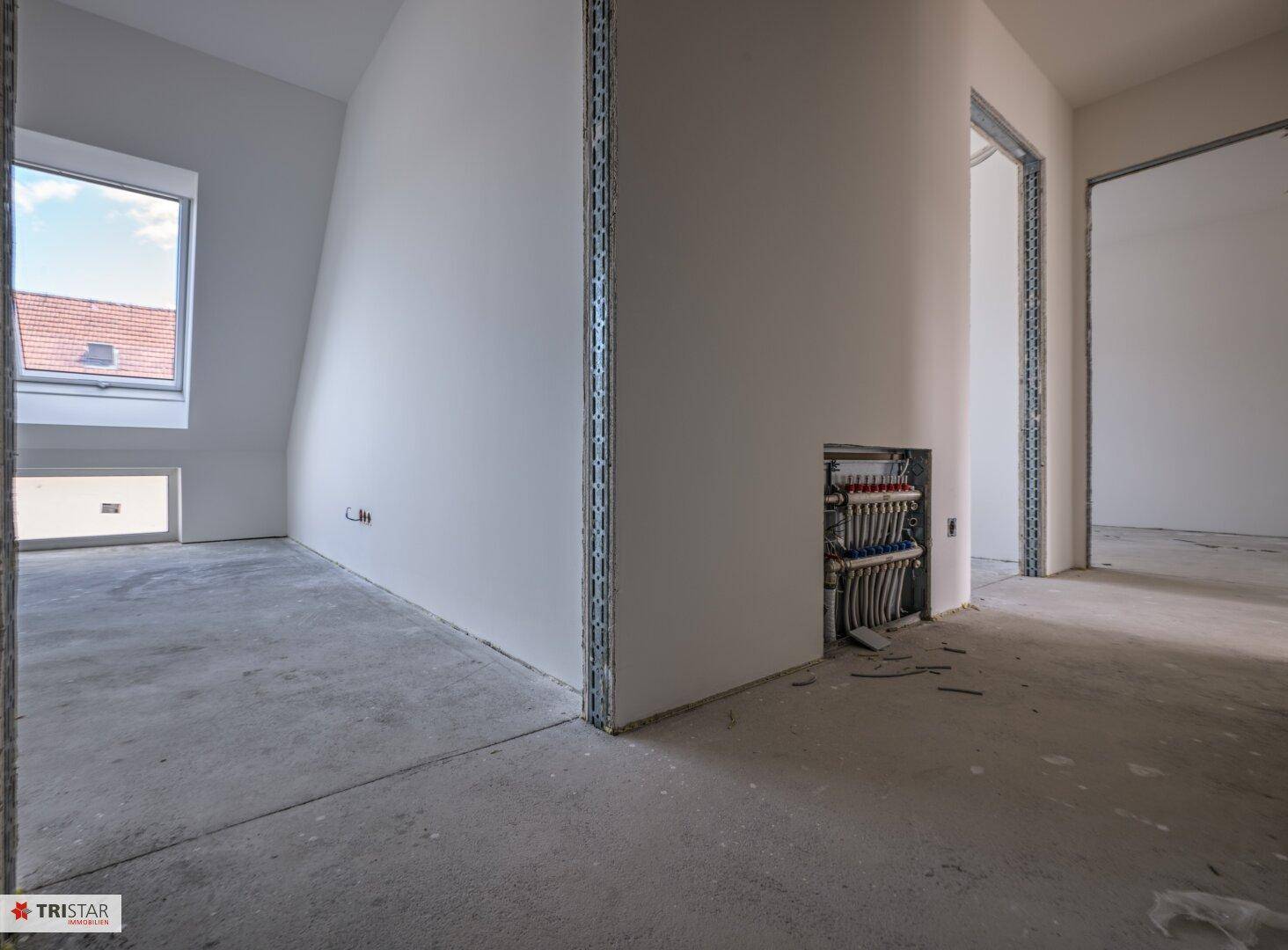 Unfertiger Dachgeschossraum mit schräger Decke, kleinem Fenster und sichtbaren Installationen in der Wand.