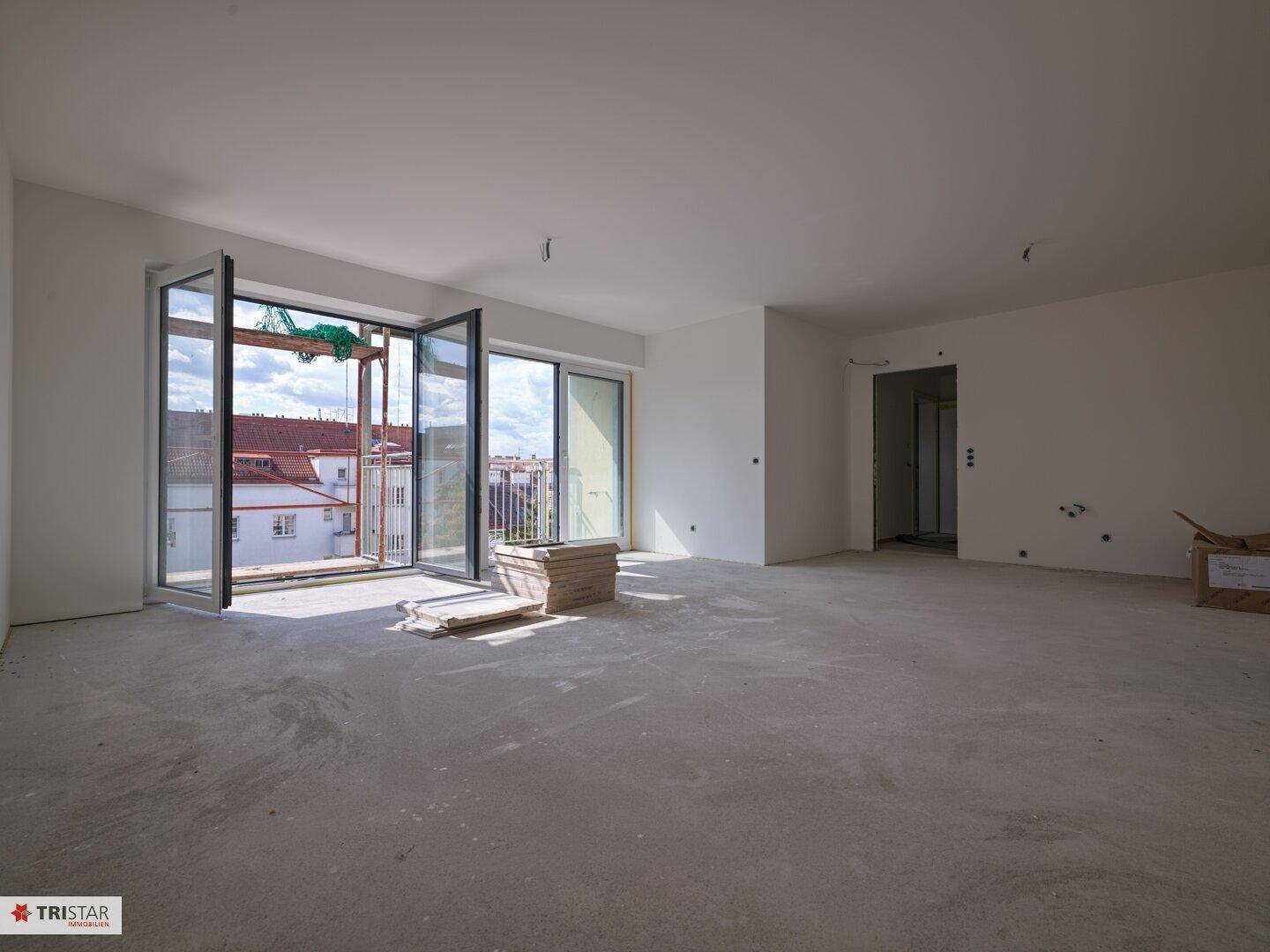 Weitläufiger, heller Wohnraum im Rohbau mit großen Fensterfronten und Betonboden.