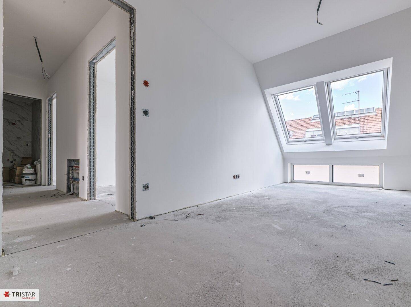 Heller Dachgeschossraum im Rohbau mit schräger Decke, Dachfenstern und Zugang zu weiteren Räumen.