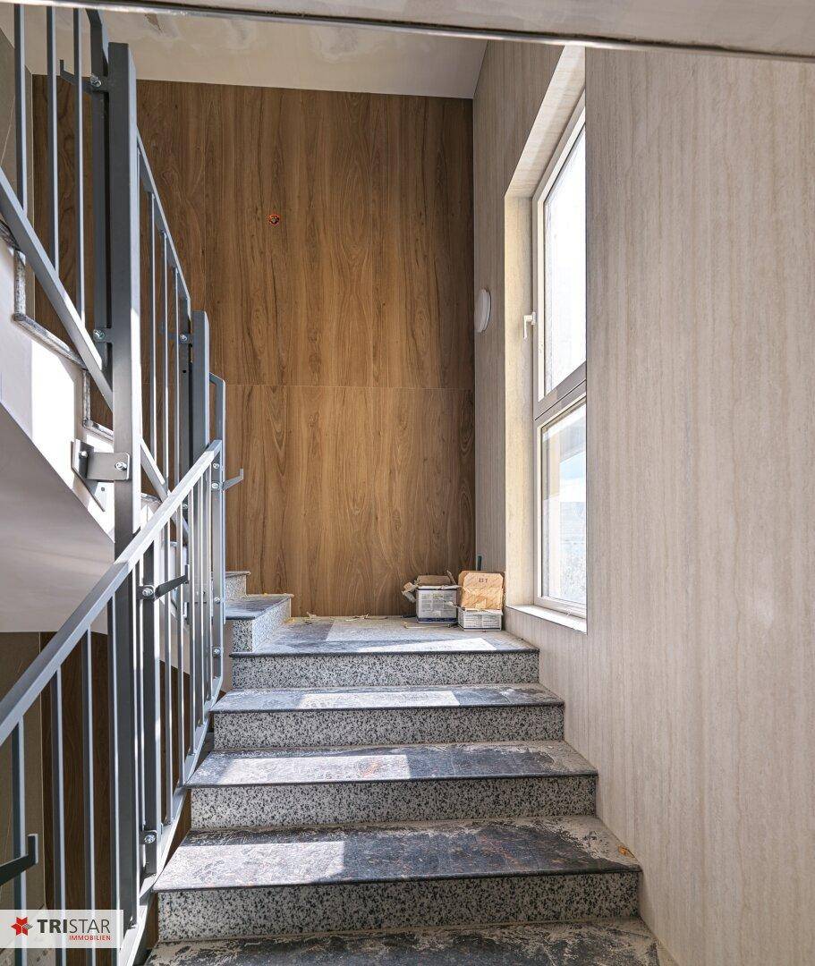 Treppenhaus im Bau mit Granitstufen, Metallgeländer und Holzverkleidung, hell durch ein Fenster.