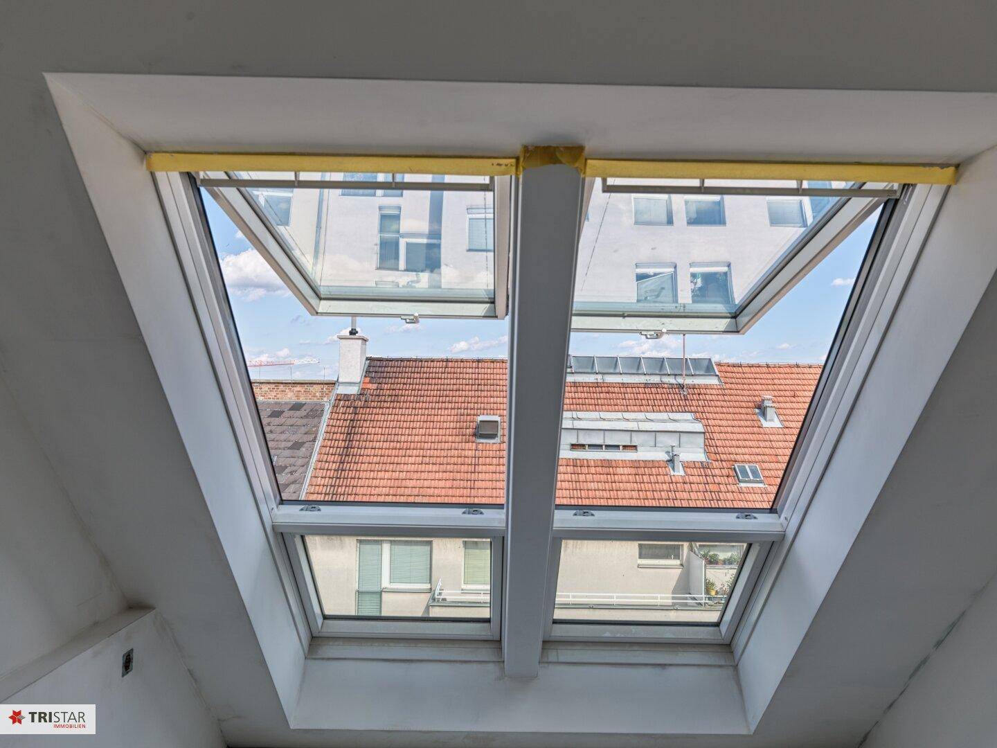 Innenraum mit zwei geöffneten Dachfenstern, die einen Blick auf die umliegenden Dächer und Gebäude freigeben.