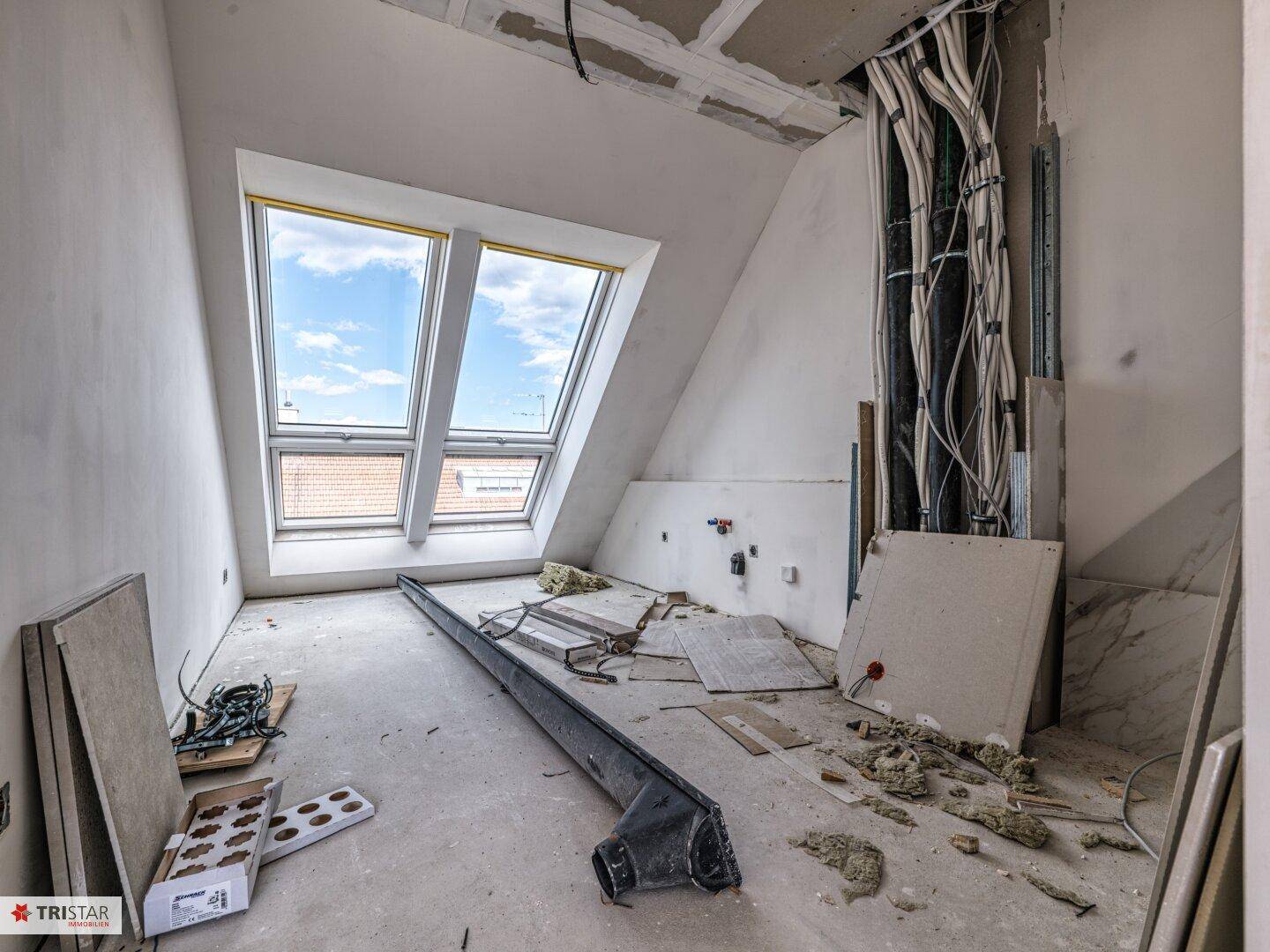 Dachgeschossraum im Rohbau mit großen Dachfenstern und sichtbaren Installationen an der Wand.