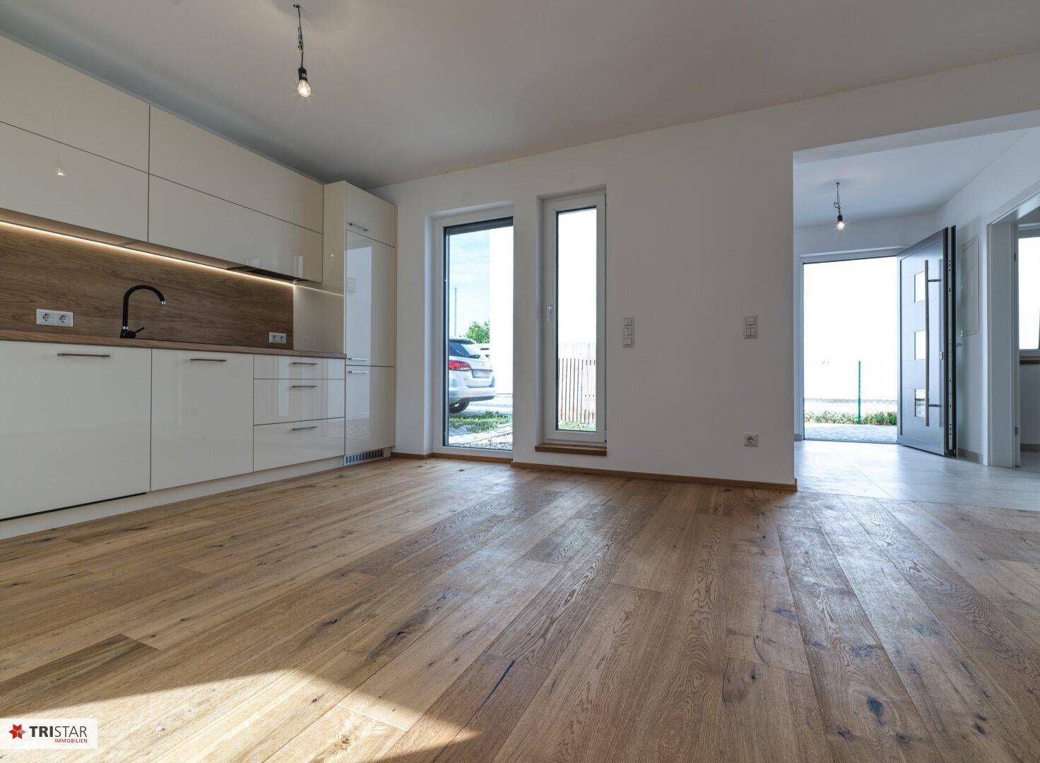 Offene Küche mit weißen Hochglanzfronten, Holzarbeitsplatte und viel Tageslicht durch große Fenster.