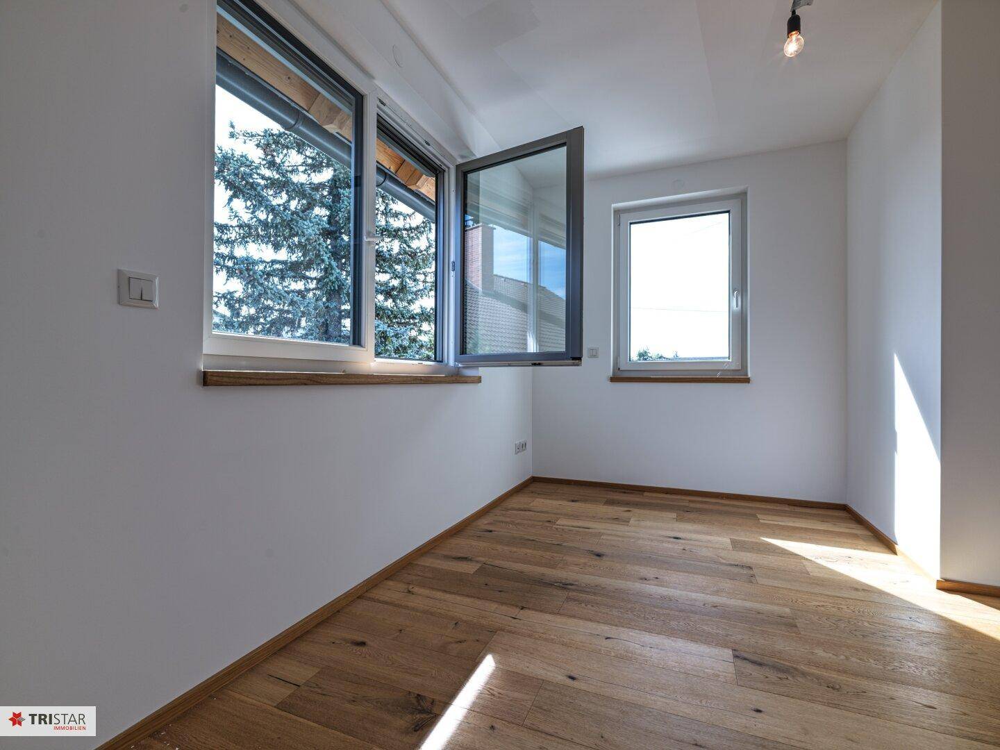 Geräumiges Zimmer mit Holzboden und großen Fenstern, die einen grünen Ausblick bieten.